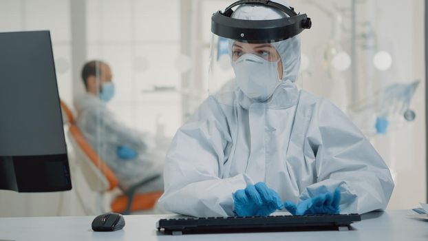 Dentistry specialist holding x ray while using computer at dental clinic desk. Caucasian nurse with ppe suit examining oral radiography scan on digital monitor for teeth healthcare