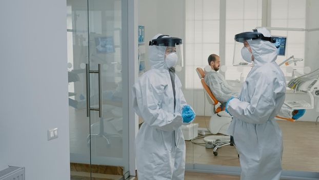 Stomatological doctors wearing ppe suits at dental clinic and standing outside cabinet discussing patient examination and operation. Caucasian dentists working during covid epidemic
