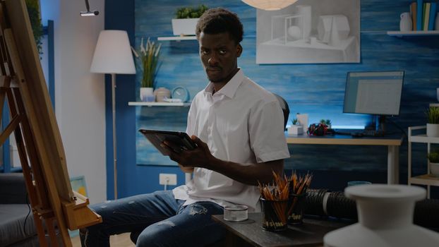 Black man with artistic skills using tablet for vase design in workshop studio. Person of african american ethnicity creating drawing on white canvas and easel with digital technology