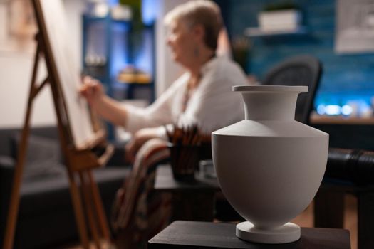 Close up of white vase on table used for authentic inspiration in workshop at studio while elder artist drawing object design on canvas in background. Senior creative person in artwork space