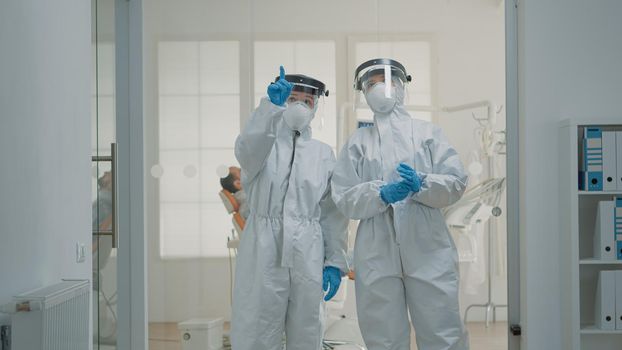 Team of caucasian dentists with hazmat suits discussing prevention for coronavirus at dentistry clinic. Professional stomatology doctors standing in dental cabinet with healthcare equipment