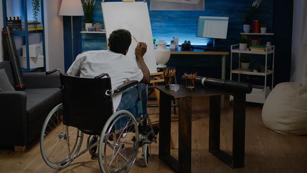 Black young artist with handicap drawing vase on canvas and easel at studio. African american creative man in wheelchair designing modern masterpiece for authentic fine art project