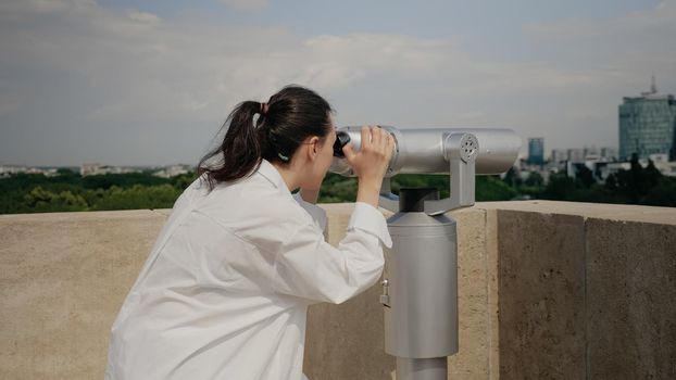 Young traveler woman using binoculars on building roof for sightseeing and urban exploration. Adult watching city lifestyle with telescope from observation point on top of skyscraper