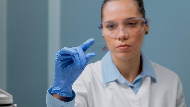 Close up of biology doctor looking at blood test on glass tray in science laboratory. Scientist woman wearing glasses and gloves for protection at clinic analyzing dna sample for experiment
