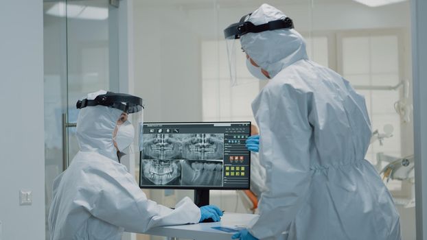 Professional dentistry staff with ppe suit looking at teeth x ray in dental cabinet. Assistant examining teeth radiography on computer monitor while doctor preparing for consultation