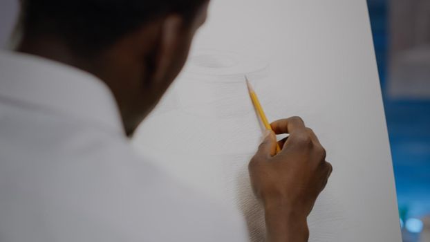 Close up of african american young artist using hand with pencil to draw vase design in workshop room. Black creative person drawing masterpiece on canvas and easel. Artistic imagination