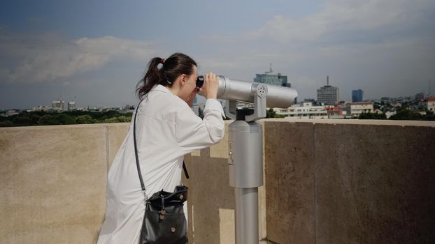 Caucasian woman using telescope from observation point on top of skyline tower in metropolis. Person with binoculars lens looking at landscape of boulevard for scenic exploration