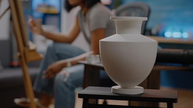 Close up of white vase used for authentic drawing project in artwork space at home. African american young artist creating masterpiece with pencil, canvas and easel in background