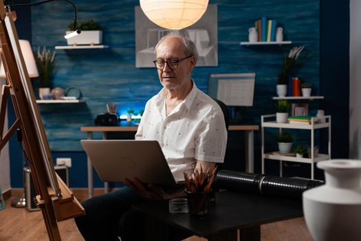 Senior artistic person using laptop for masterpiece drawing on canvas in workshop studio at home. Caucasian elderly artist with gadget creating fine art for professional artwork hobby