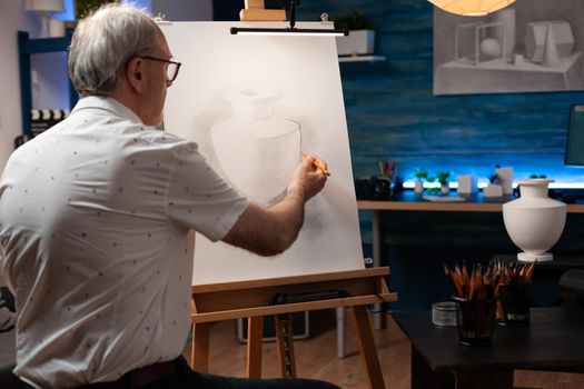 Senior artist using vase for drawing inspiration on canvas in workshop space. Caucasian elder man working on authentic masterpiece with professional instruments in artwork studio