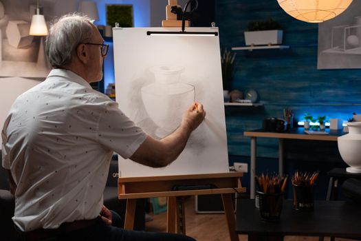 Elderly artist working on vase drawing with pencil on canvas and easel at art studio. Caucasian senior man recreating authentic masterpiece with object for fine art project. Old creative person