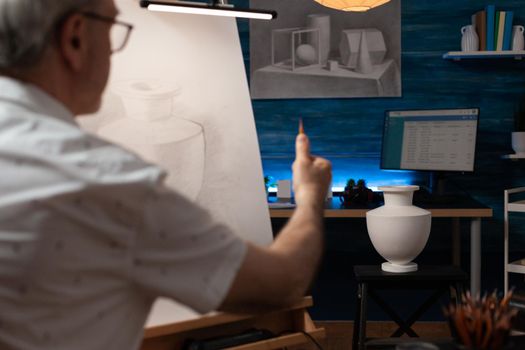 Caucasian senior man drawing vase design on white canvas in fine art studio. Elderly artist creating professional masterpiece on wooden easel in artwork space. Old adult with artistic hobby