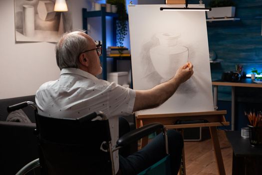 Senior artist with disability drawing vase design with pencil in artwork room at home. Caucasian man sitting in wheelchair creating masterpiece on white canvas and easel