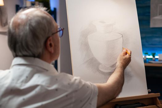 Caucasian elder artist working on vase masterpiece in artistic workspace. Senior man sitting in wheelchair drawing using pencil on white canvas with wooden easel in fine art studio