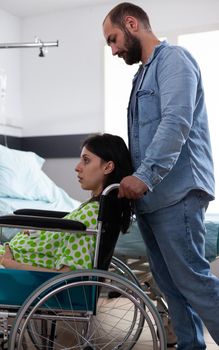 Caucasian man helping woman with pregnancy in wheelchair at medical clinic. Husband supporting pregnant wife while sitting in hospital ward with professional healthcare equipment