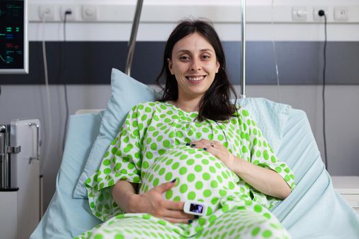 Portrait of woman with pregnancy smiling and holding hand on belly while looking at camera in hospital ward. Caucasian adult expecting baby and sitting in bed preparing for childbirth