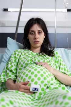 Portrait of tired pregnant person sitting in hospital ward bed at medical facility. Woman expecting child, looking at camera and holding hands on belly. Young adult with pregnancy