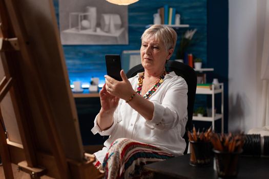Senior artist using smartphone sitting in art studio room for pictures of drawing masterpiece. Old woman looking at modern cellphone for inspiration indoors. Adult with technology for fine art