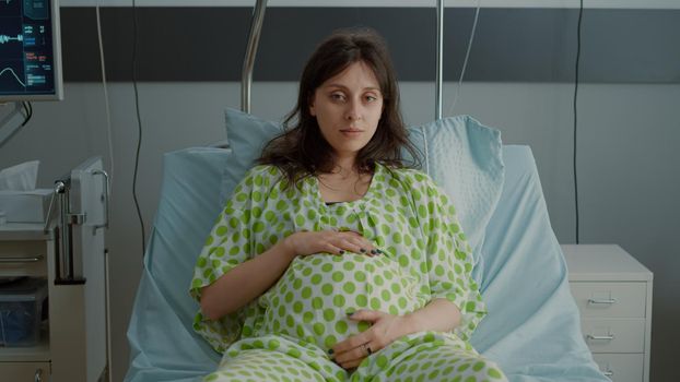 Portrait of pregnant woman laying in hospital ward bed with medical equipment preparing for childbirth at maternity. Caucasian patient holding hand on belly while sitting expecting baby