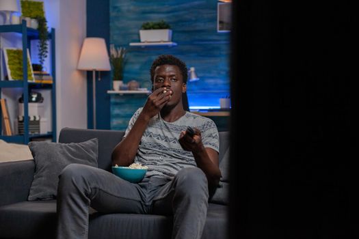 African american young man sitting on couch changing channel using remote while eating popcorn spending free time alone in living room. Guy watching entertainment movie on television