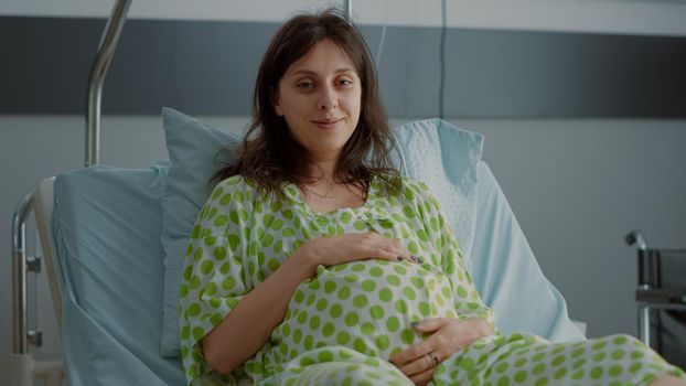 Portrait of adult with pregnancy holding belly in hospital ward waiting for assistance and support. Caucasian young woman laying in bed expecting baby delivery. Pregnant patient