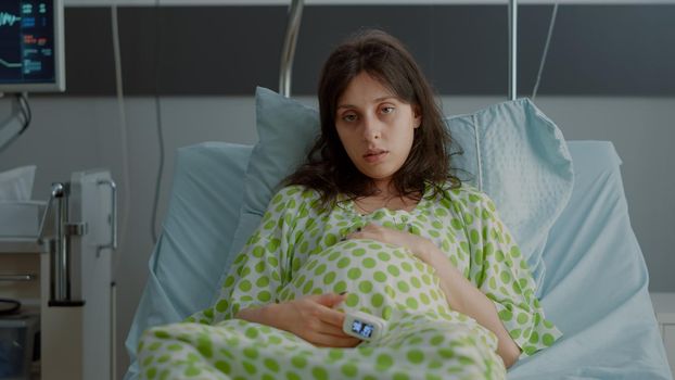 Tired caucasian woman with pregnancy belly sitting in bed at maternity room. Young pregnant person with oximeter feeling tension about childbirth in hospital ward. Adult preparing for delivery