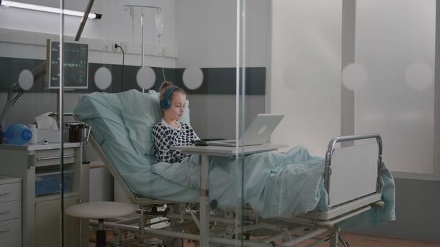 Alone sick girl patient with oxygen nasal tube relaxing in bed wearing headphones while watching entertainment movie on laptop computer during medical examination. Girl recovery after surgery