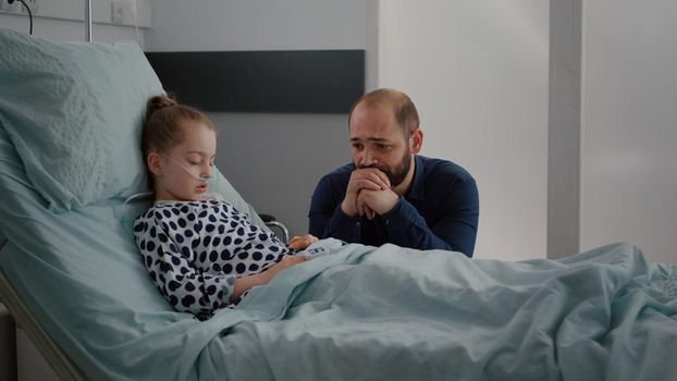 Sick child daughter wearing oxygen nasal tube and oximeter on finger monitoring heartbeat pulse while sleeping. Worried father crying beside bed during recovery period in hospital ward, medical treatment