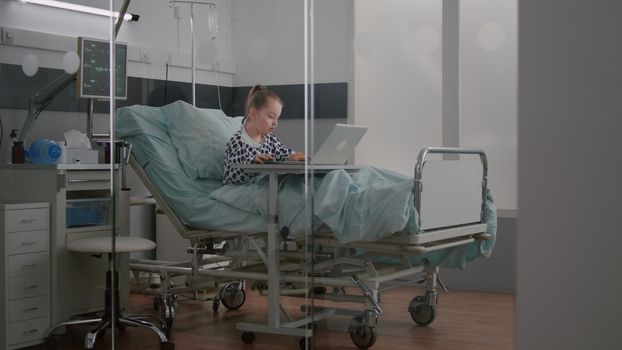 Sick child relaxing in bed playing cartoon video games on laptop computer during medical examination in hospital ward. Hospitalized kid having breathing sickness recovering after surgery