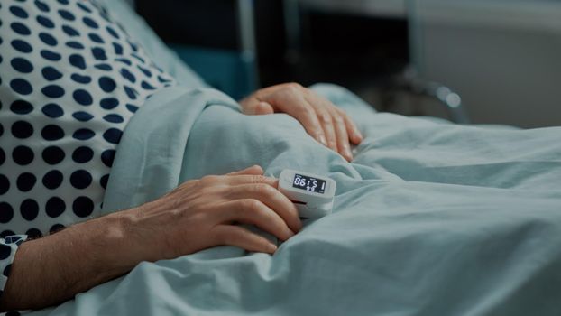 Close up of oximeter on patient in hospital ward bed at medical facility. Old man waiting on results for treatment against health diagnosis. Doctor checking IV drip bag with serum