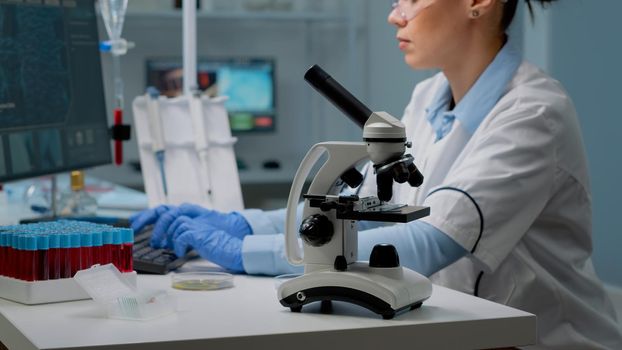 Close up of microscope tool while scientist analyzing petri dish and using modern computer in background. Woman at laboratory desk with blood sample and tray of vacutainers for examination