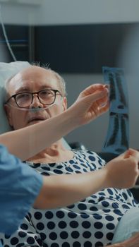 Medical nurse explaining xray results to sick patient in hospital ward at recovery clinic. Elder man with IV drip bag and nasal oxygen tube laying in bed with serious health problems