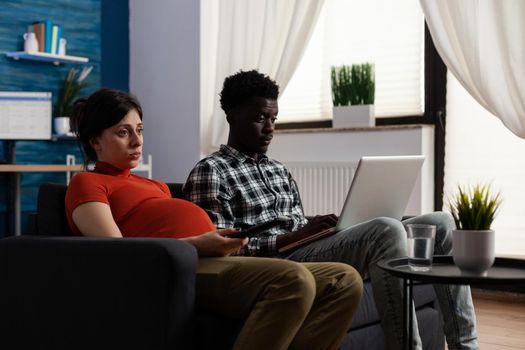 Interracial couple expecting child and using technology at home. African american man working on digital laptop while pregnant caucasian woman laying on sofa. Mixed race partners with pregnancy