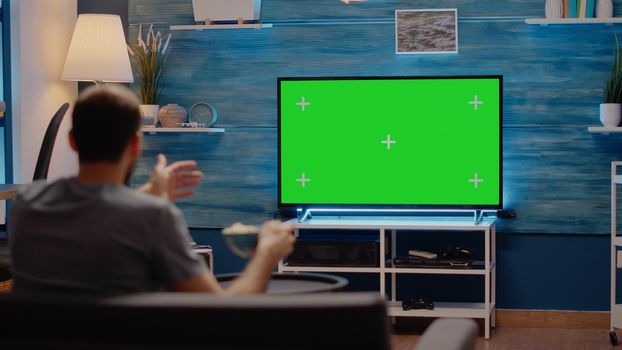 Person watching sport event or football on green screen television cheering in living room at modern home using isolated media display. Chroma key background for blank mockup template