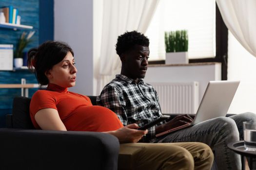 Interracial couple expecting child and using technology at home. African american man working on modern laptop while pregnant caucasian woman switching channels with TV remote control.