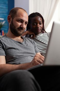 Close up of interracial couple looking at laptop screen at home. Caucasian man working on digital device and african american woman watching display. Multi ethnic people with technology