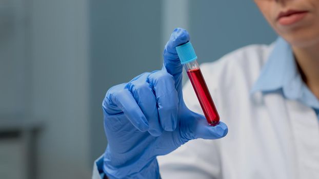 Close up of vacutainer filled with blood for investigation in laboratory. Scientist with gloves looking at transparent glassware tool with liquid solution used by doctor for science