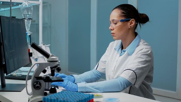 Researching specialist working in laboratory with chemical equipment. Woman using computer for dna examination while having microscope, petri dish, vacutainers and micropipette on desk