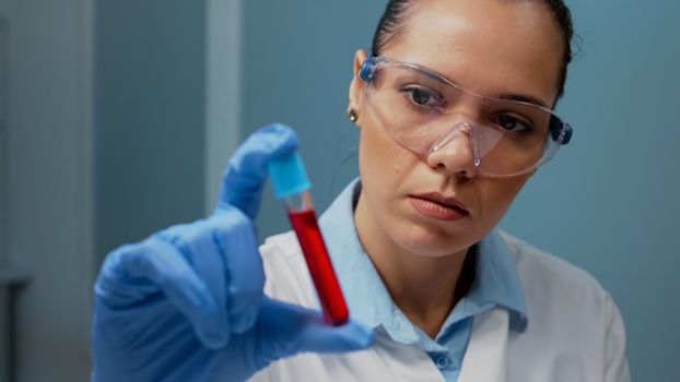 Laboratory scientist holding vacutainer with blood sample in chemical workplace. Chemist wearing gloves and glasses looking at transparent tube with red substance for medical development