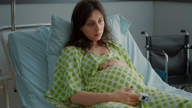 Portrait of pregnant adult holding hand on belly in hospital ward waiting for pregnancy healthcare. Caucasian woman sitting in bed with tension expecting baby. Childbirth young patient