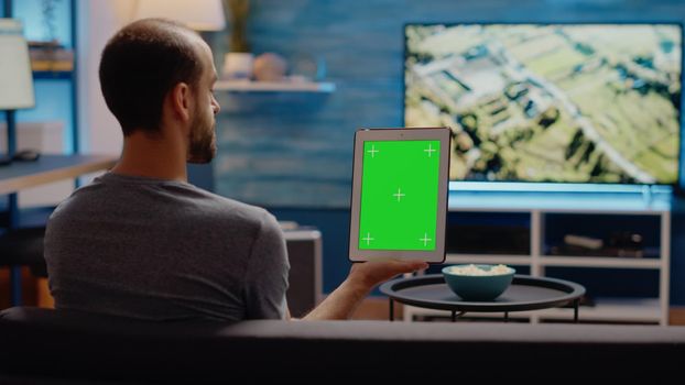 Young man vertically holding digital tablet with green screen design in living room. Modern technology for chroma key used on mockup template and isolated display for copy space