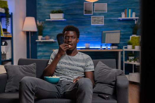 African american guy watching entertainment movie on television relaxing on sofa at home. Black young man looking at camera while eating popcorn snack enjoying spending free time alone in living room