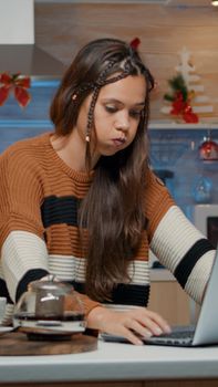 Festive adult typing on laptop while sitting in kitchen decorated with christmas ornaments and tree. Young woman looking at watch and waiting for friends preparing for holiday dinner