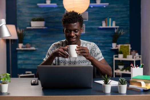 African american businessman holding cup of coffee typing management ideas on computer working remote at online project in living room. Freelancer sitting at desk analyzing financial graph