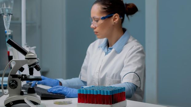 Microbiology specialist using computer in laboratory while having vacutainers tray with fluid blood samples and microscope prepared for genetic examination. Chemist woman in lab coat