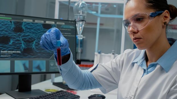 Doctor in science laboratory analyzing tray of vacutainers on desk. Chemistry scientist looking at lab glassware tube filled with fluid blood for medicine development. Woman wearing gloves