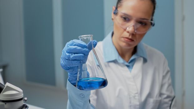 Portrait of microbiology doctor analyzing chemical beaker in laboratory. Scientific woman using lab glassware for analysis. Chemist holding flask with blue solution for medical development