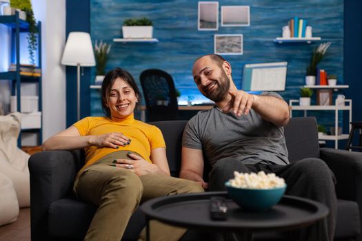 Caucasian married couple expecting child while relaxing at home. Young pregnant woman smiling with hand on baby bump while man pointing at television, looking at camera and watching TV