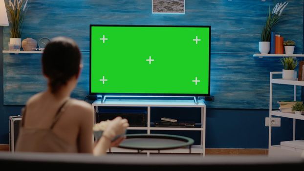 Woman looking at tv designed with green screen mockup template and isolated blank background. Young person sitting in living room at home using chroma key on display and copy space