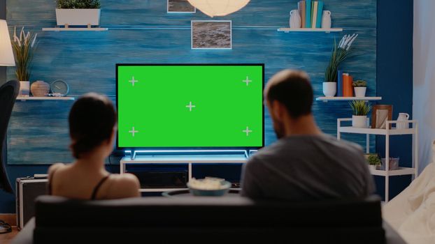 Caucasian individuals using television for green screen sitting on couch in living room. People with chroma key for virtual layout and mockup isolated template. Copy space background